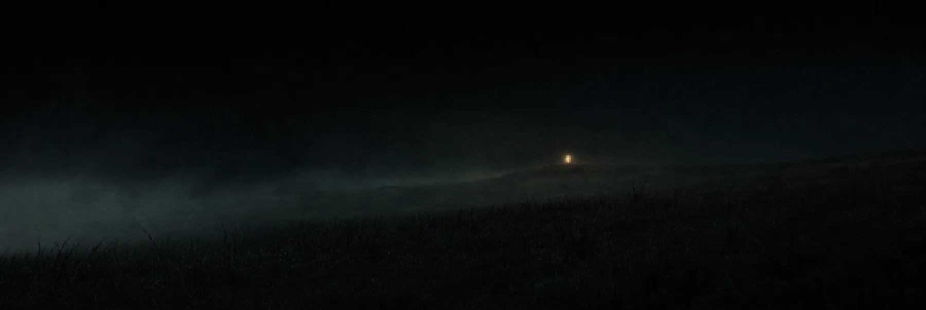 A dark hillside field at night with fog and a single small lamp burning far off in the distance.