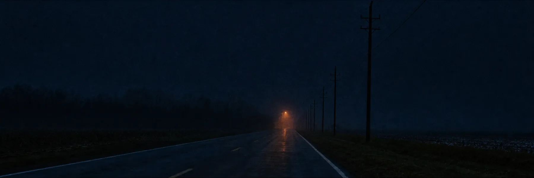 An empty two-lane road at blue hour, one distant amber streetlight glowing at the horizon.